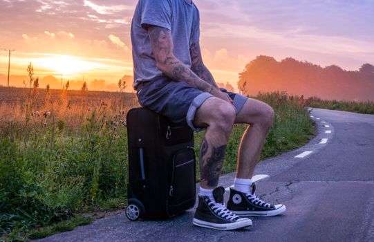 A tattooed individual sits on luggage by a country road during sunrise, wearing casual attire.