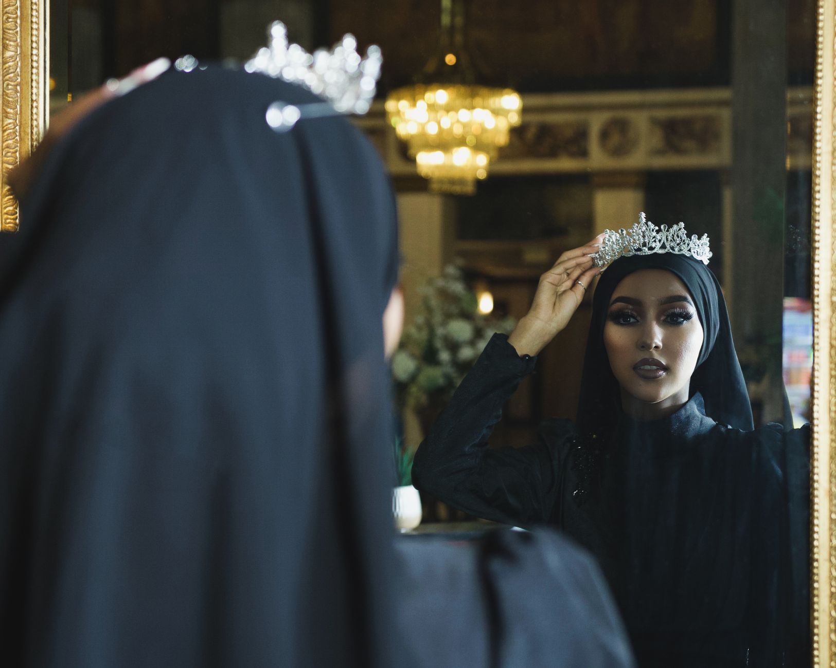 Muslim woman in hijab admires herself wearing a crown in front of a mirror.