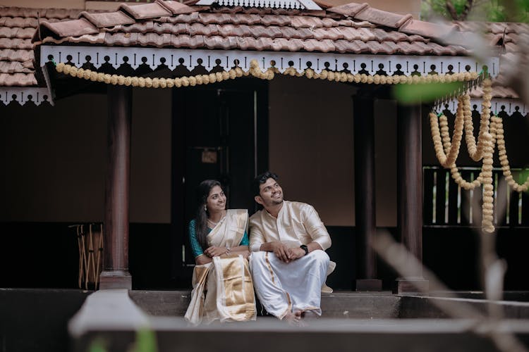 Smiling Couple In Traditional Clothes Sitting On Stairs Near House