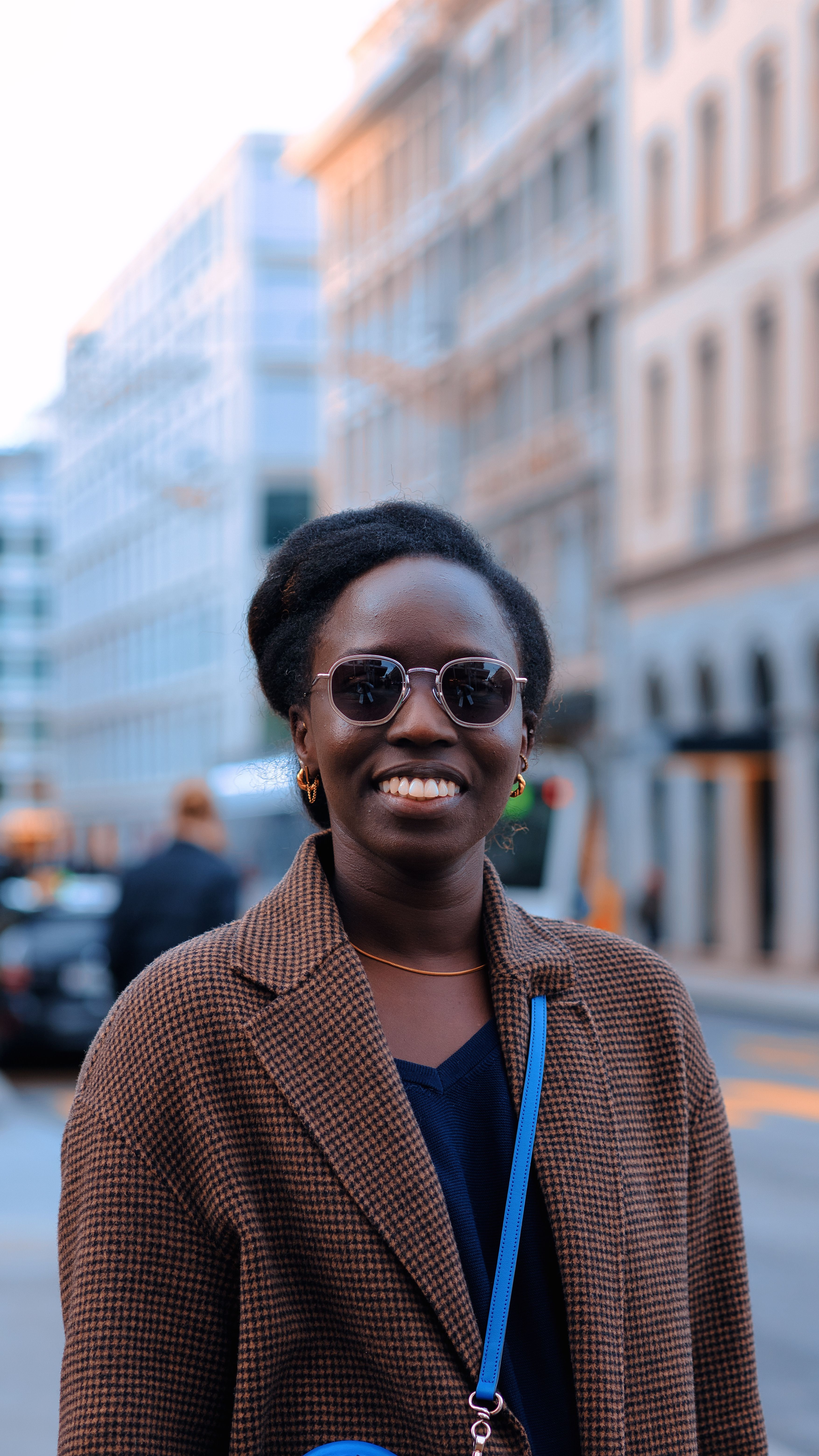 Elegant woman in a coat smiling on a city street with tall buildings and a lively urban backdrop.