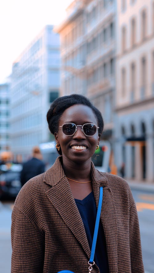 Young Woman in a Coat and Sunglasses Standing on the Street and Smiling 