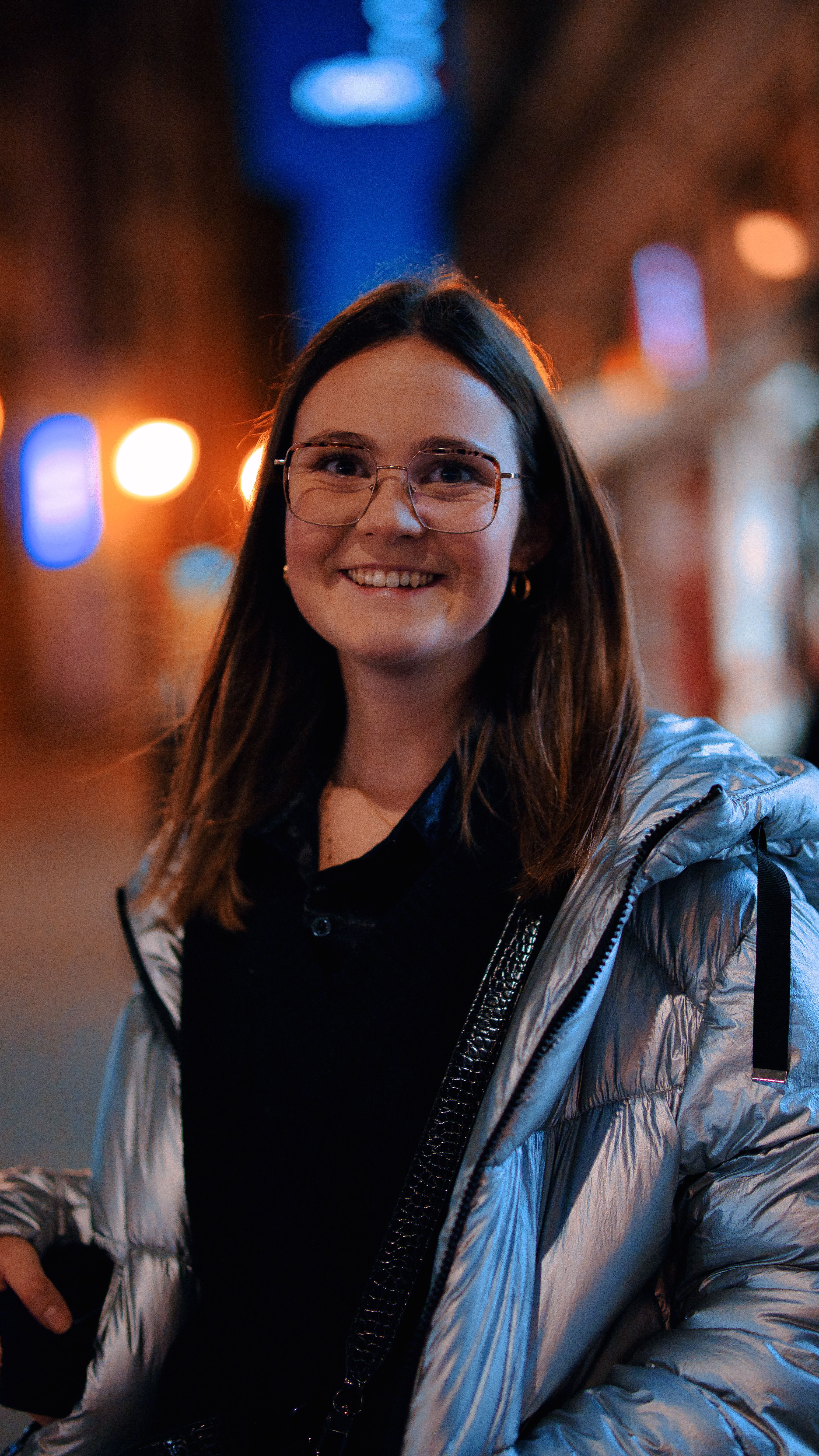 A happy woman with eyeglasses and a shiny jacket smiling in a well-lit urban street at night.