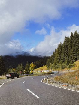 Curving mountain road with a car, surrounded by pine trees and cloudy skies.