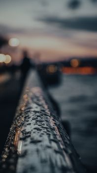 A close-up of a wet railing by the waterfront at sunset, with blurred city lights in the background.