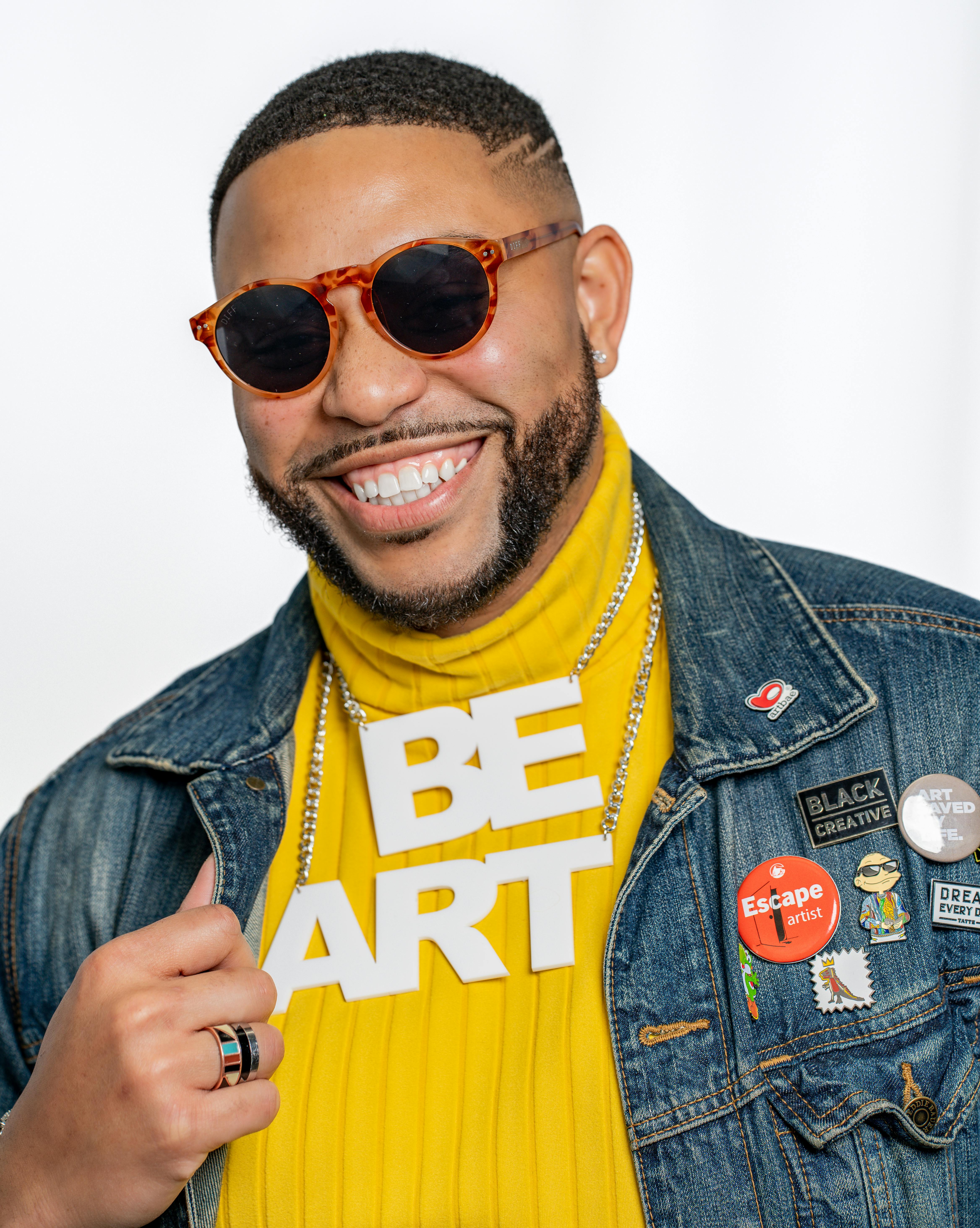 Smiling man in sunglasses and denim jacket with artistic pins on a white background.
