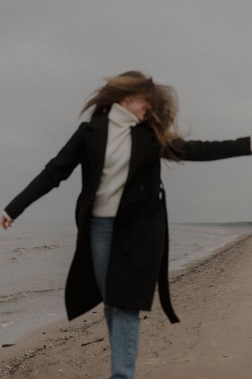 Free Stylish woman in a black coat dancing freely on a breezy beach day. Stock Photo