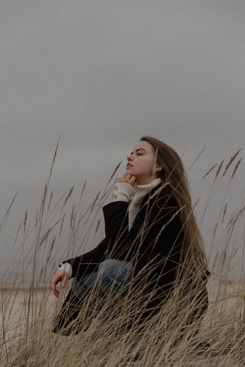Free Woman in winter coat sits in tall grass, contemplating the serene snowy landscape. Stock Photo
