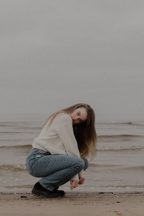 Free A young woman in casual attire squats pensively near the seaside on a cloudy day. Stock Photo