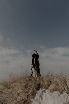 A woman with her dog stands on a grassy hill under a clear blue sky.