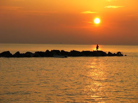 A lone fisherman silhouettes against a vibrant sunset, casting from a rocky outcrop by the sea.