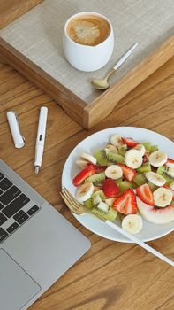 A delicious fruit salad and coffee on a wooden desk beside a laptop, perfect for a work break.