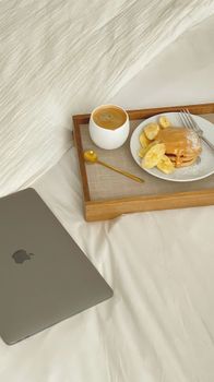 Tray with coffee and pancakes on bed, laptop nearby for cozy morning setup.