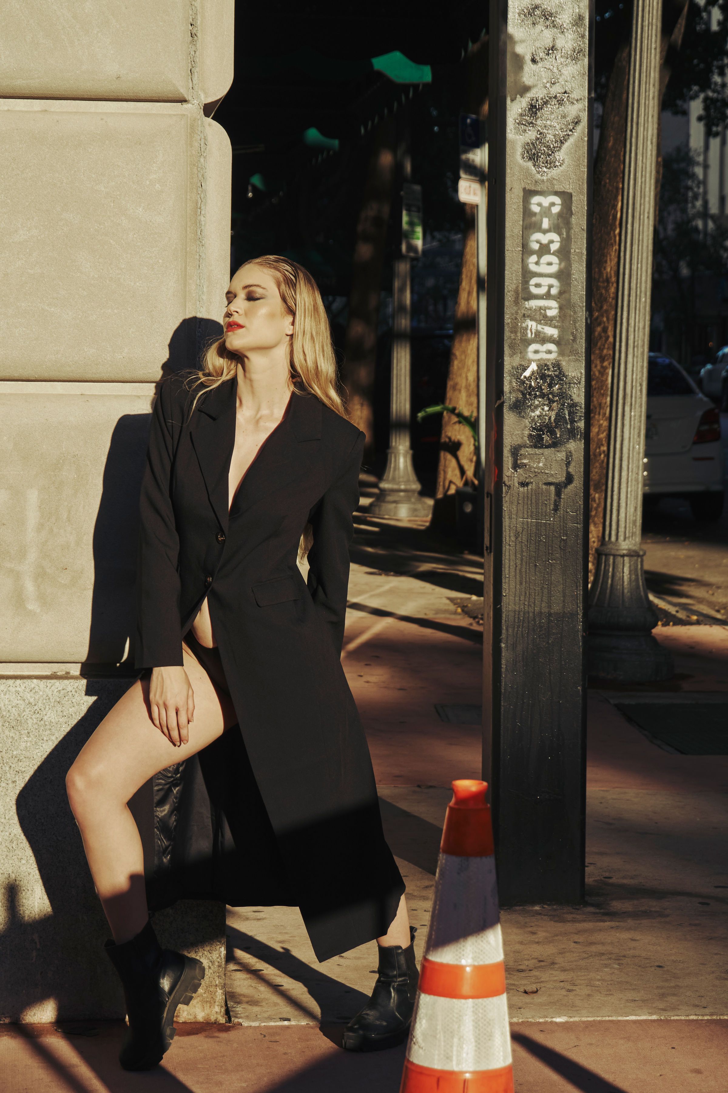 Stylish woman in a black coat posing in a sunlit city street, exuding elegance and allure.