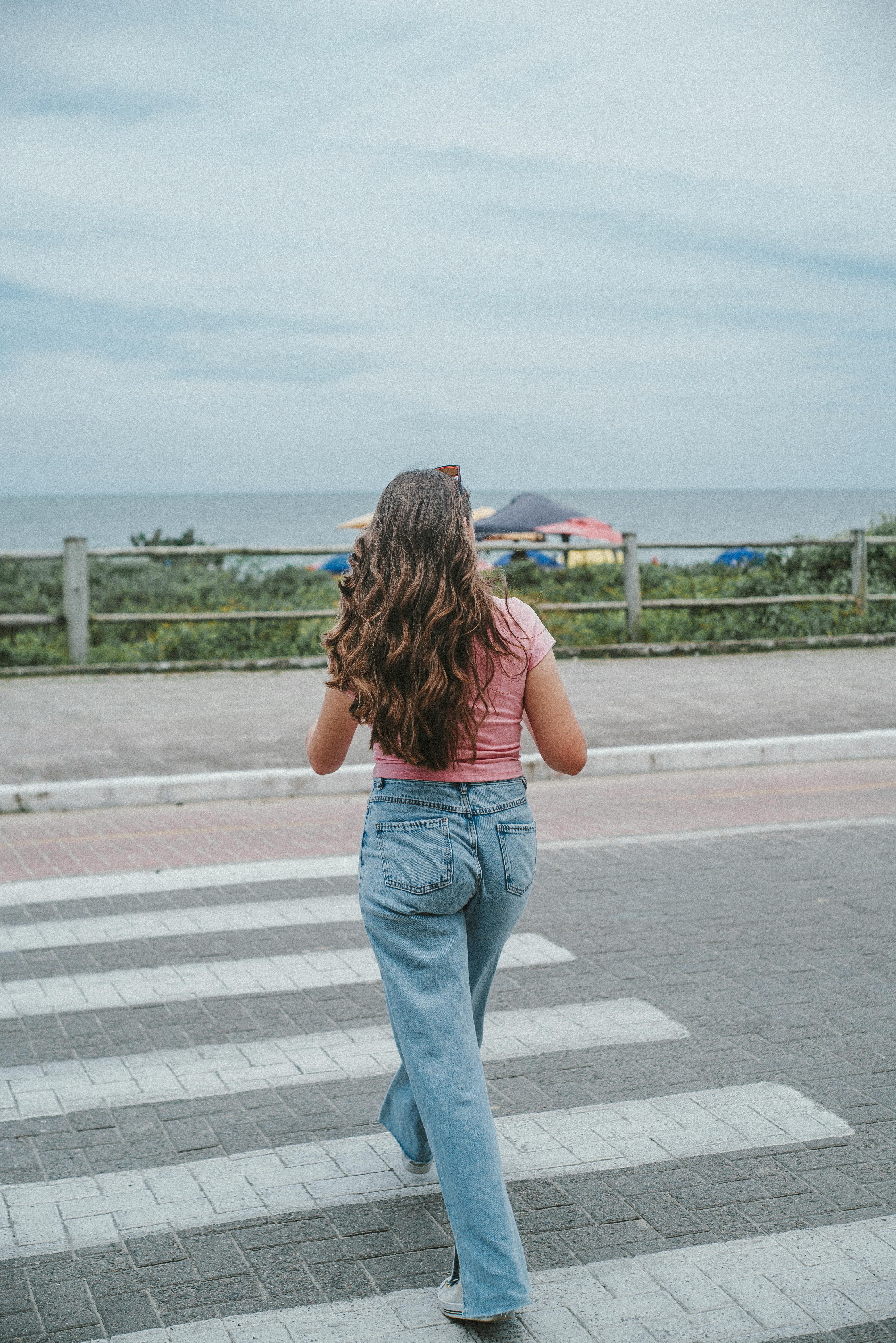 Woman with long hair walking on a crosswalk by the beach in Itajaí, Brazil, wearing a pink top and jeans.