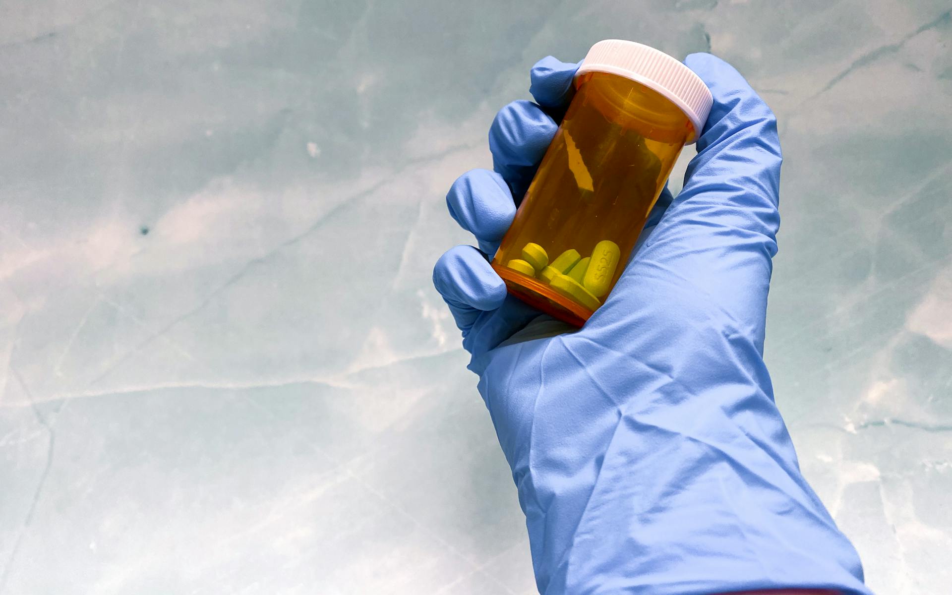 Close-up of a gloved hand holding an orange prescription pill bottle with pills inside on a gray background.