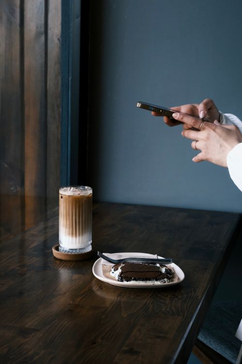 Free A person photographs coffee and cake on a wooden table in a stylish café setting. Stock Photo