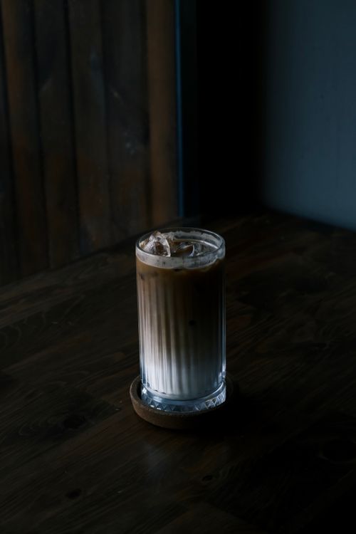 Free A tall glass of iced coffee sits on a wooden table in a dimly lit café environment. Stock Photo