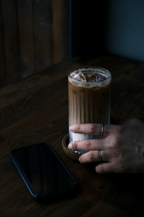 Free A hand with rings holds a glass of iced coffee next to a smartphone on a wooden table. Stock Photo