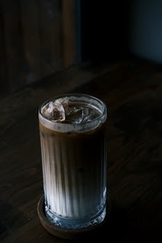 A glass of iced latte with ice cubes on a wooden table, captured in a dimly lit café.