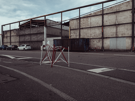 A deserted city street in an industrial area featuring cars, container, and concrete structures.
