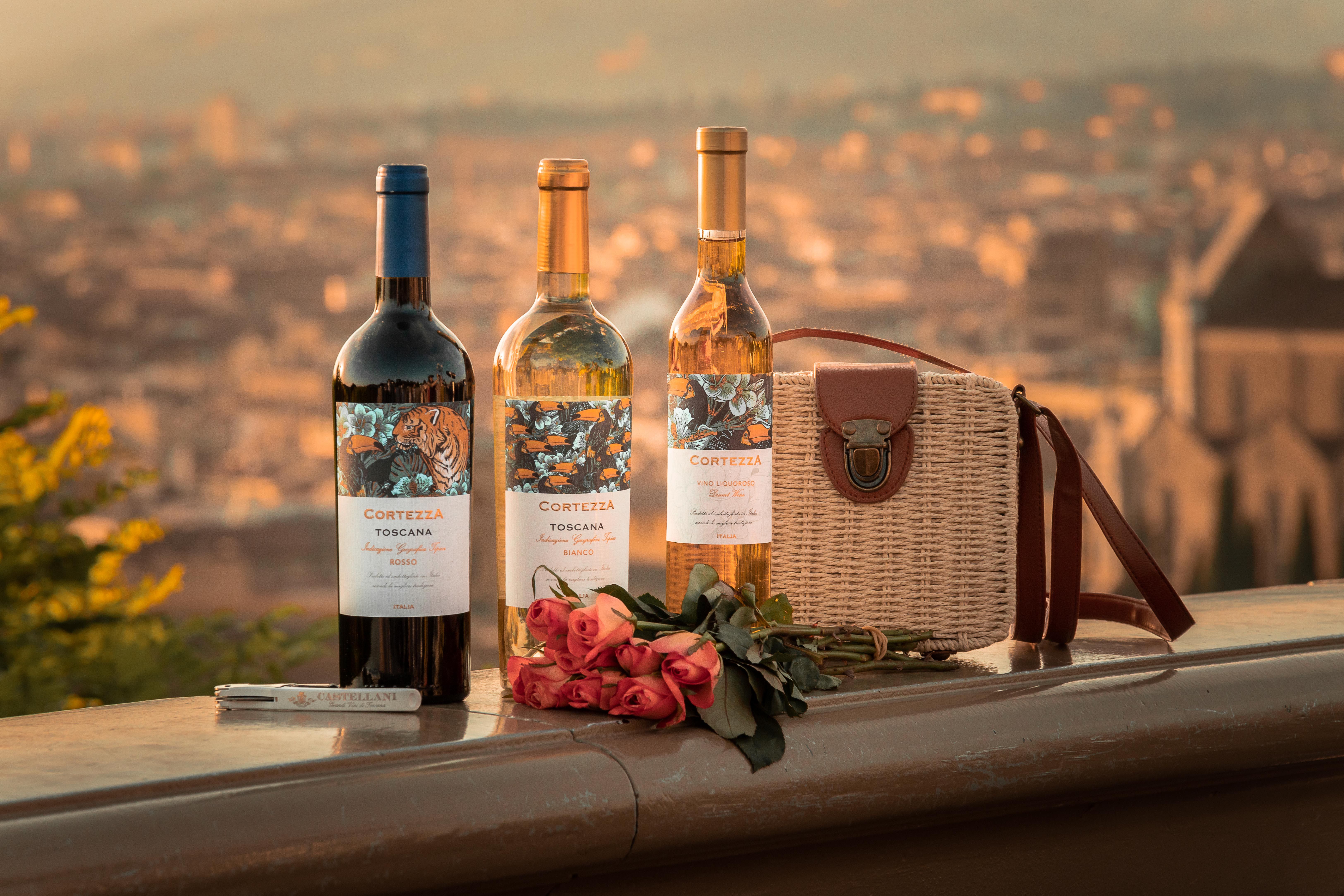 Wine bottles and roses with a Tuscan town backdrop create a romantic scene.