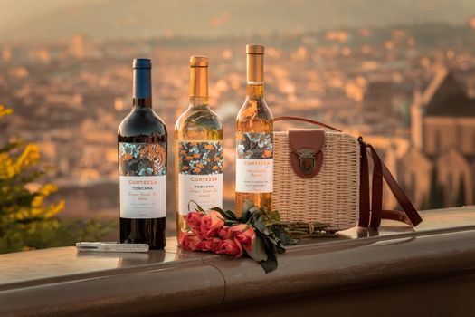 Wine bottles and roses with a Tuscan town backdrop create a romantic scene.