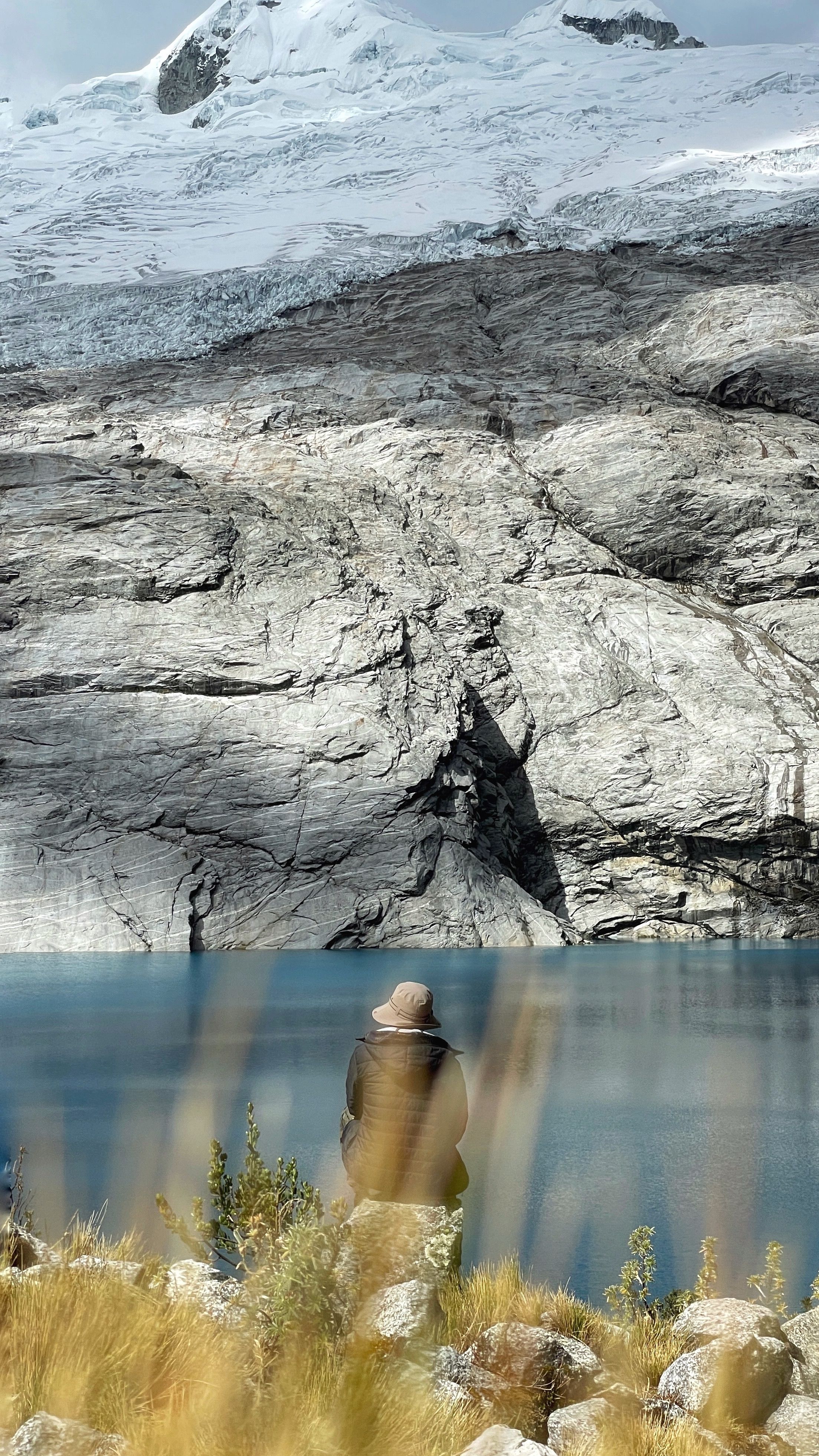 A lone person sits by a tranquil lake under majestic snowcapped mountains, offering a sense of peace and solitude.
