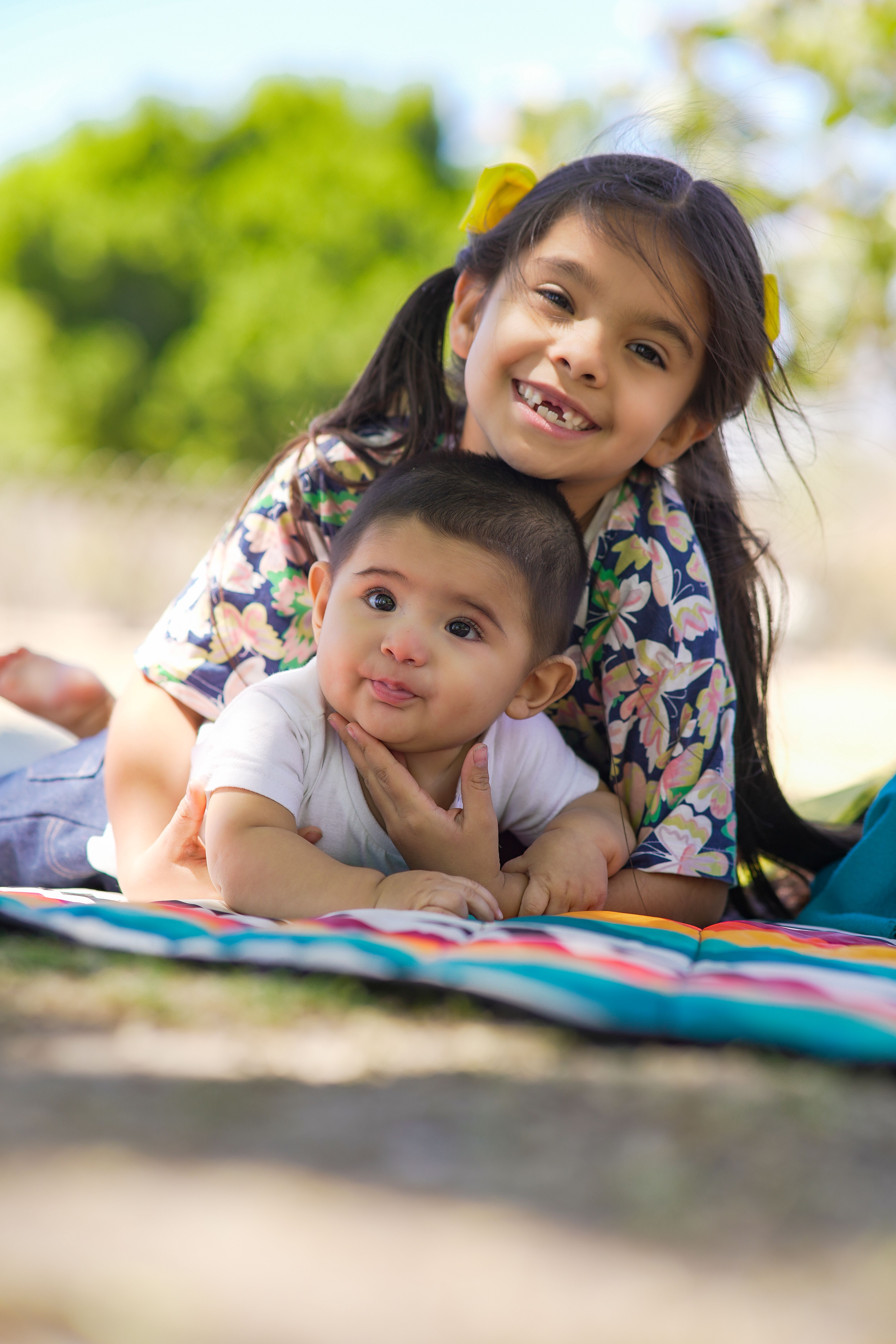 Fotomural Dos hermanos sonrientes, una niña y un bebé, disfrutando de un día soleado al aire libre sobre una manta colorida.