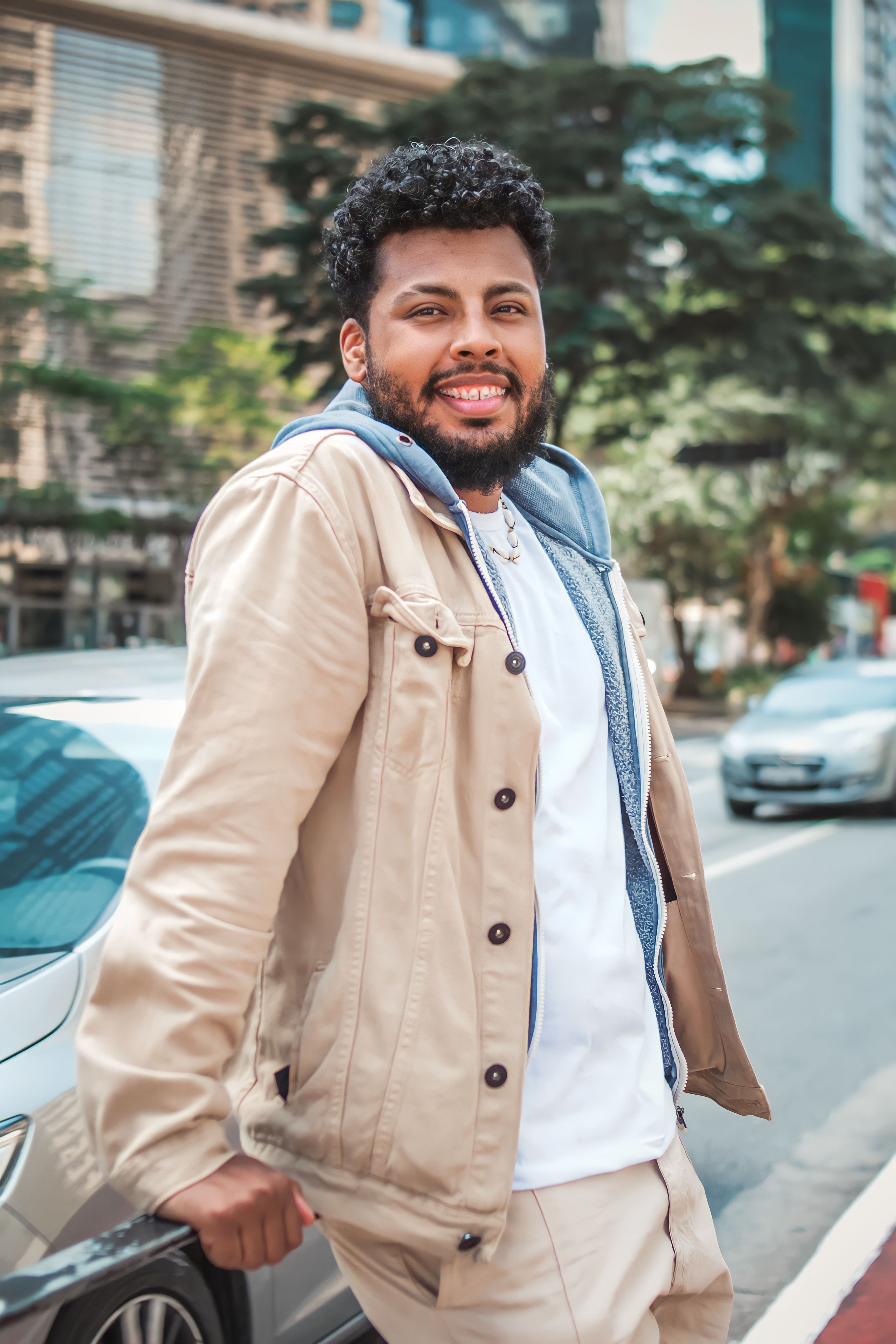 A young man in a casual outfit smiling while leaning on a car in the city.