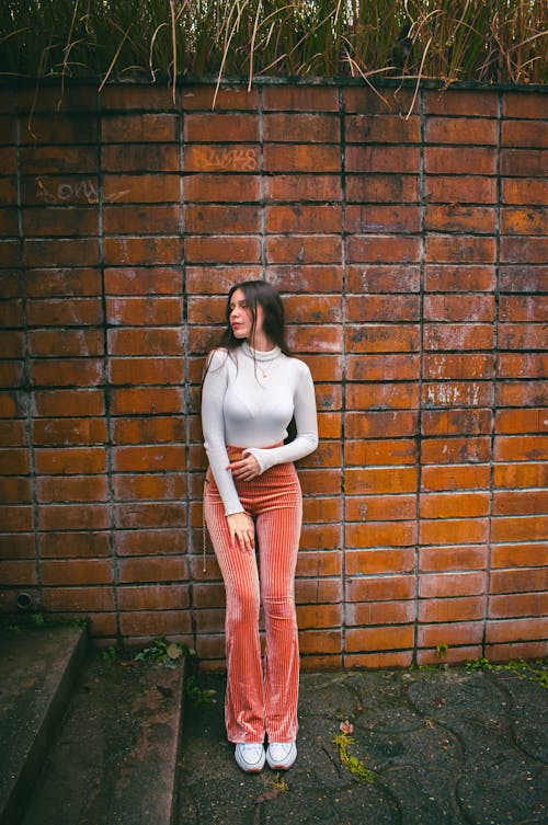 Young Woman in a White Blouse and Pink Trousers Standing against a Brick Wall 