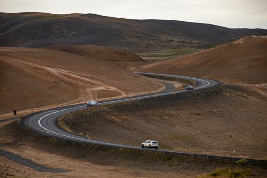 Cars drive along a winding road through a barren desert landscape under cloudy skies.