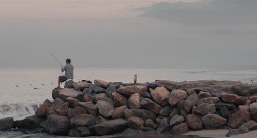 Back View of a Man Sitting on a Rocky Shore and Fishing 