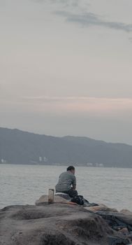A man sits quietly on rocks, gazing across a peaceful bay under a cloudy sky.