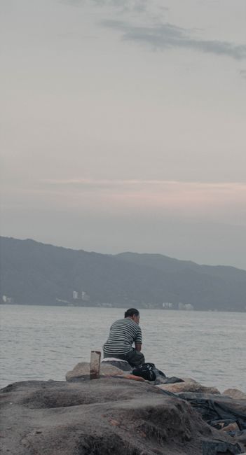 Fotomural Un hombre se sienta tranquilamente en las rocas, mirando hacia una bahía pacífica bajo un cielo nublado.