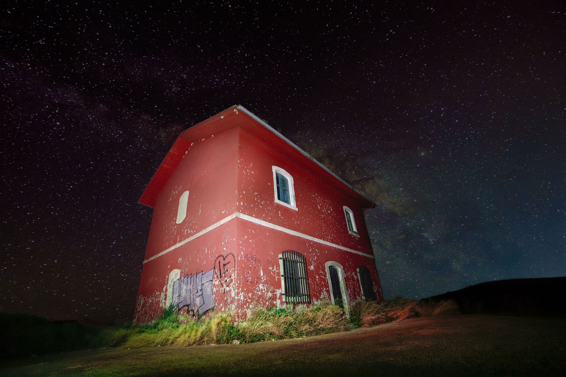 A red abandoned house stands against a vibrant starry night sky, showcasing stunning celestial views.