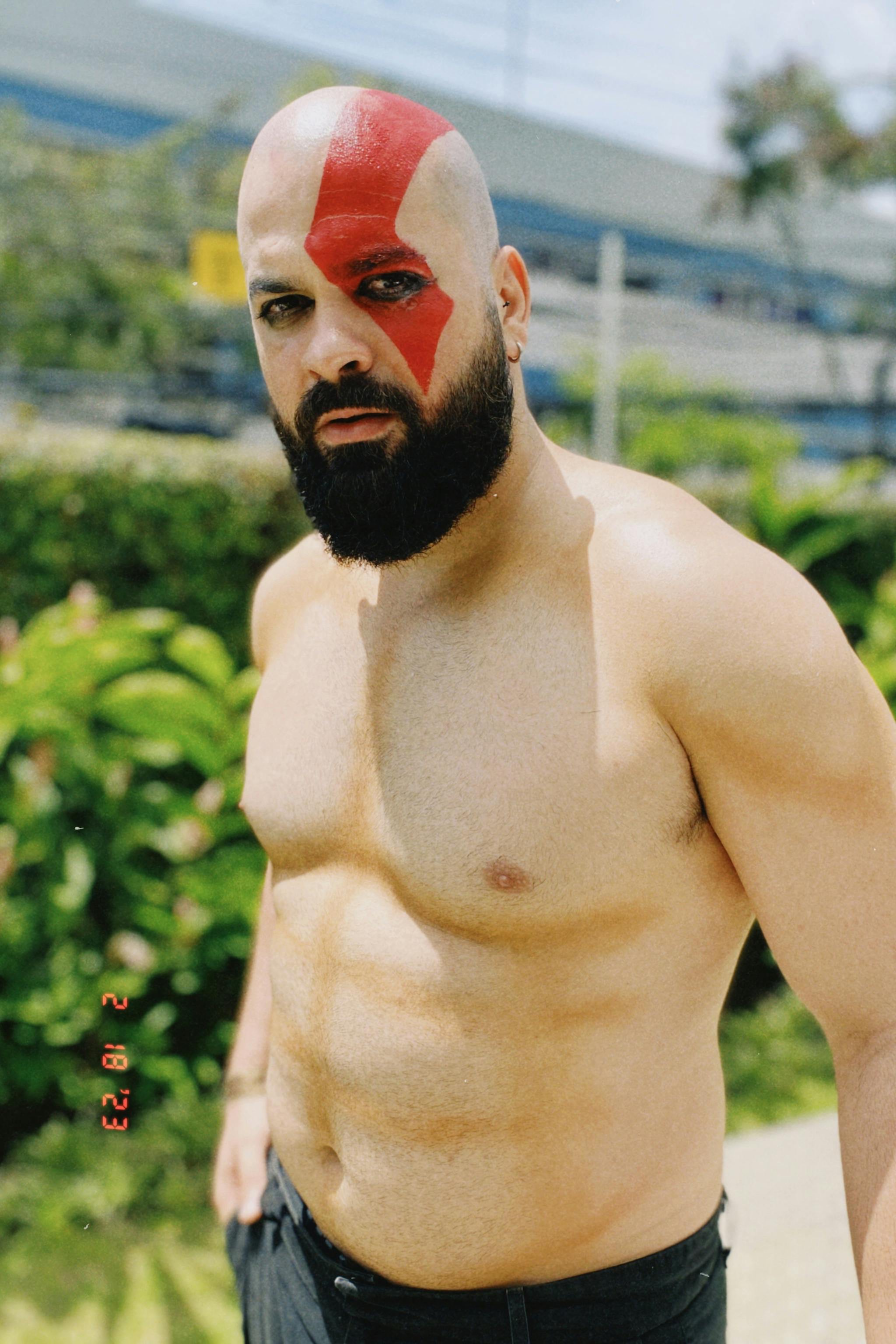 A muscular bald man with a beard and face paint standing outdoors, looking at the camera.