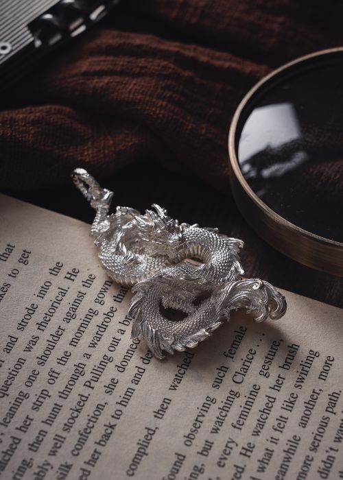 Free Artistic silver dragon placed on vintage book with magnifying glass detail. Stock Photo