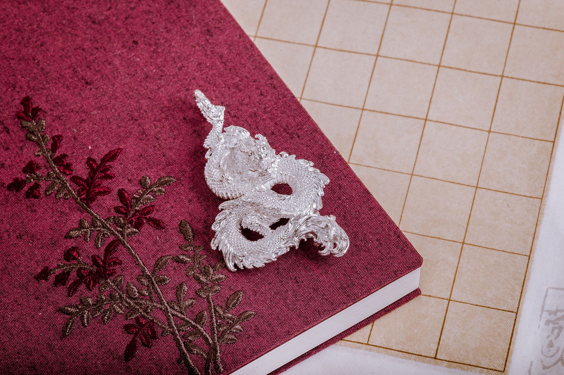 Silver dragon ornament placed on a detailed embroidered maroon journal with a vintage aesthetic.