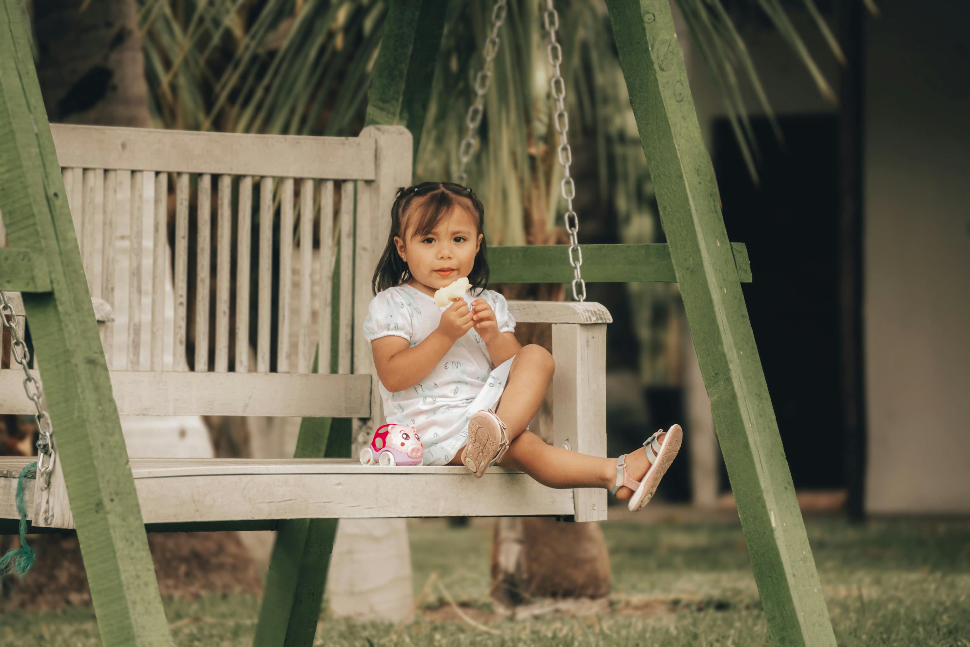 A young girl enjoying ice cream on a wooden swing outdoors with a toy nearby.