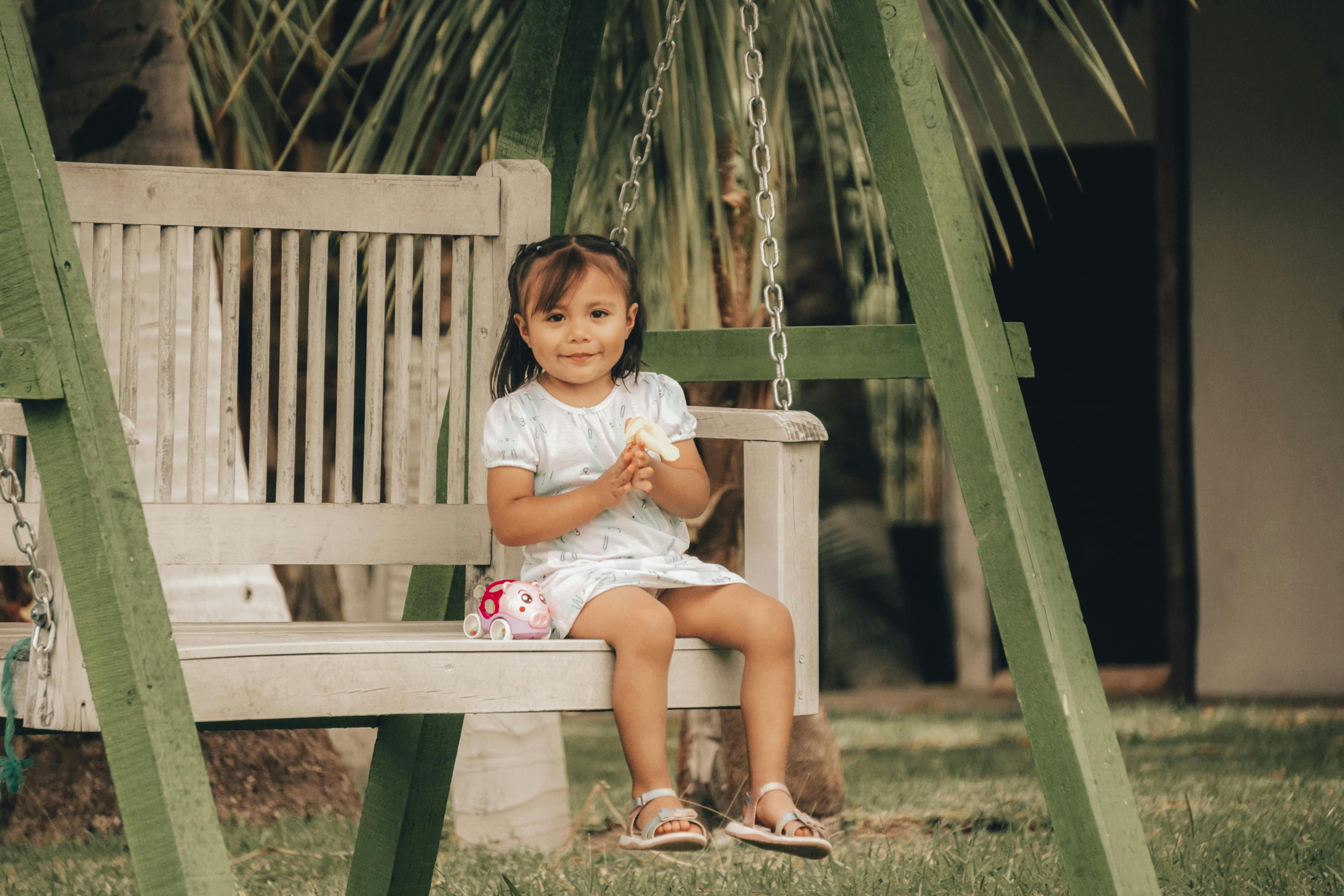 Cute brunette girl in a dress sitting on a wooden swing, smiling outdoors.