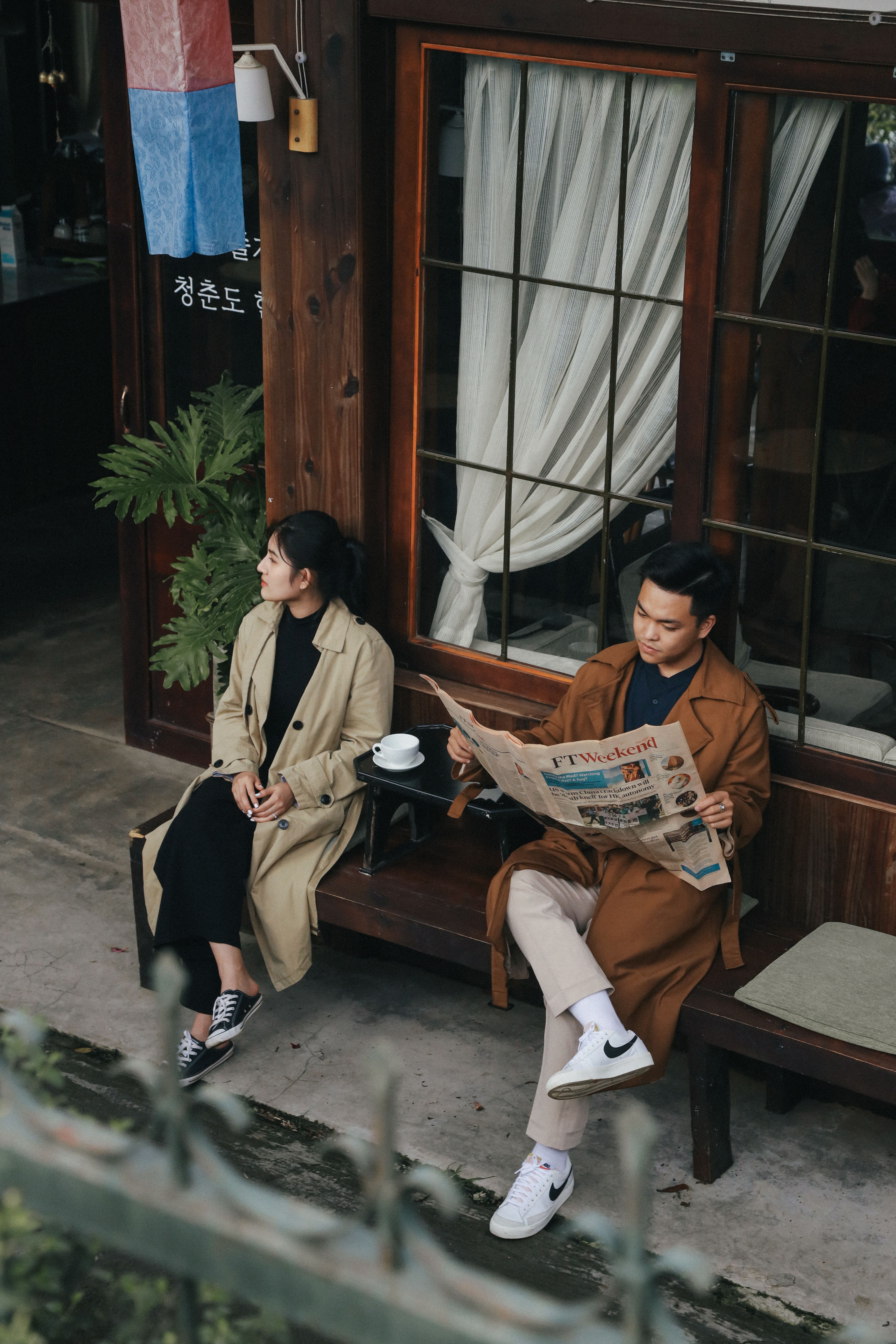 A man and woman in coats are sitting outdoors at a cafe, with the man reading a newspaper.