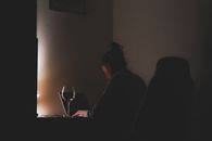 A woman working on a laptop with a glass of wine in a dimly lit room.