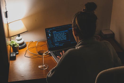 A person working from home at night, typing on a laptop with soft lamp lighting and a glass of wine.