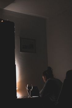 A woman working on a laptop in a dimly lit room, with a glass of wine on the desk, creating a moody atmosphere.