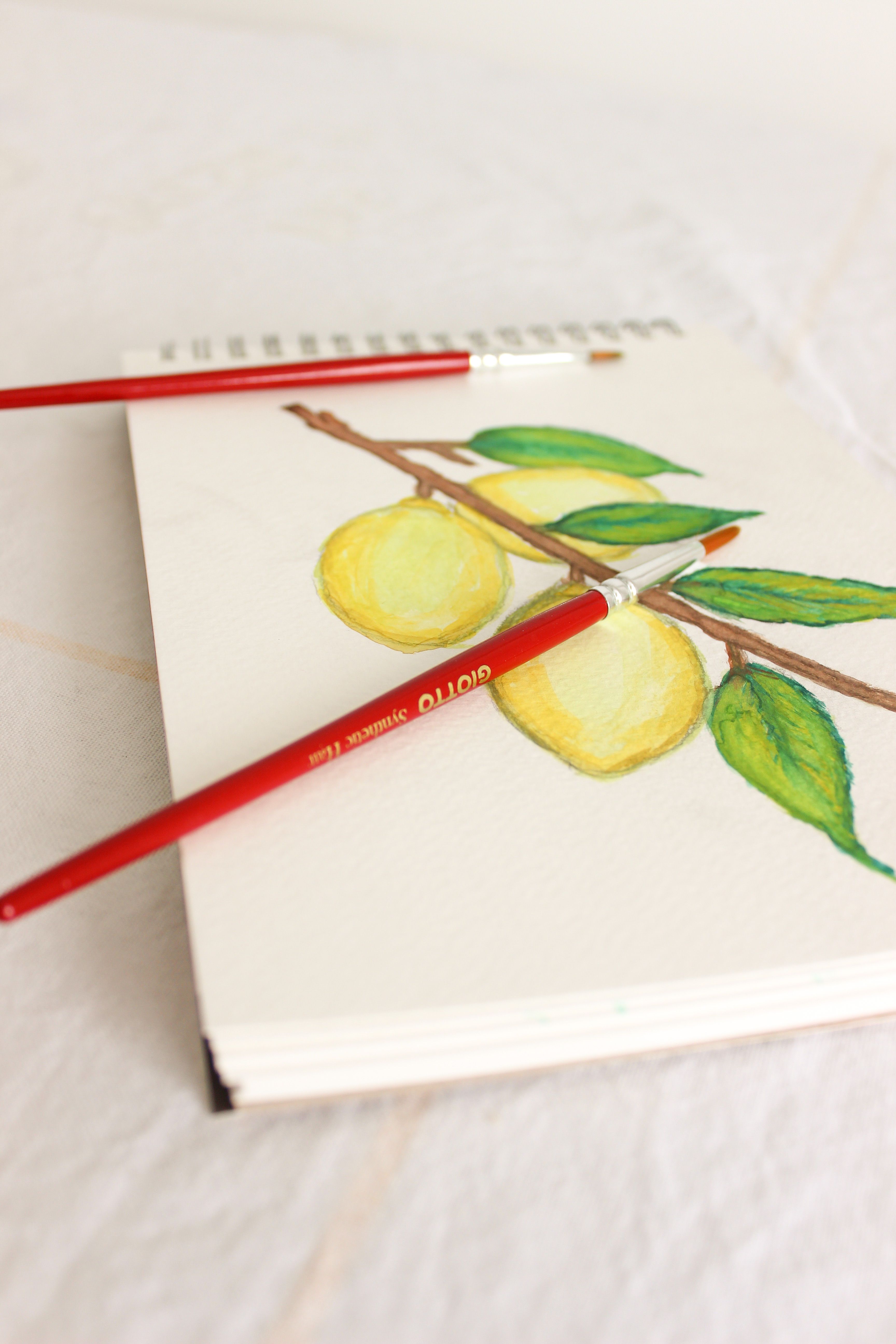 Vibrant lemon illustration in a sketchbook with two paintbrushes on paper.