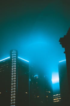 Tall buildings in a misty urban setting, illuminated by cool blue lights at night.