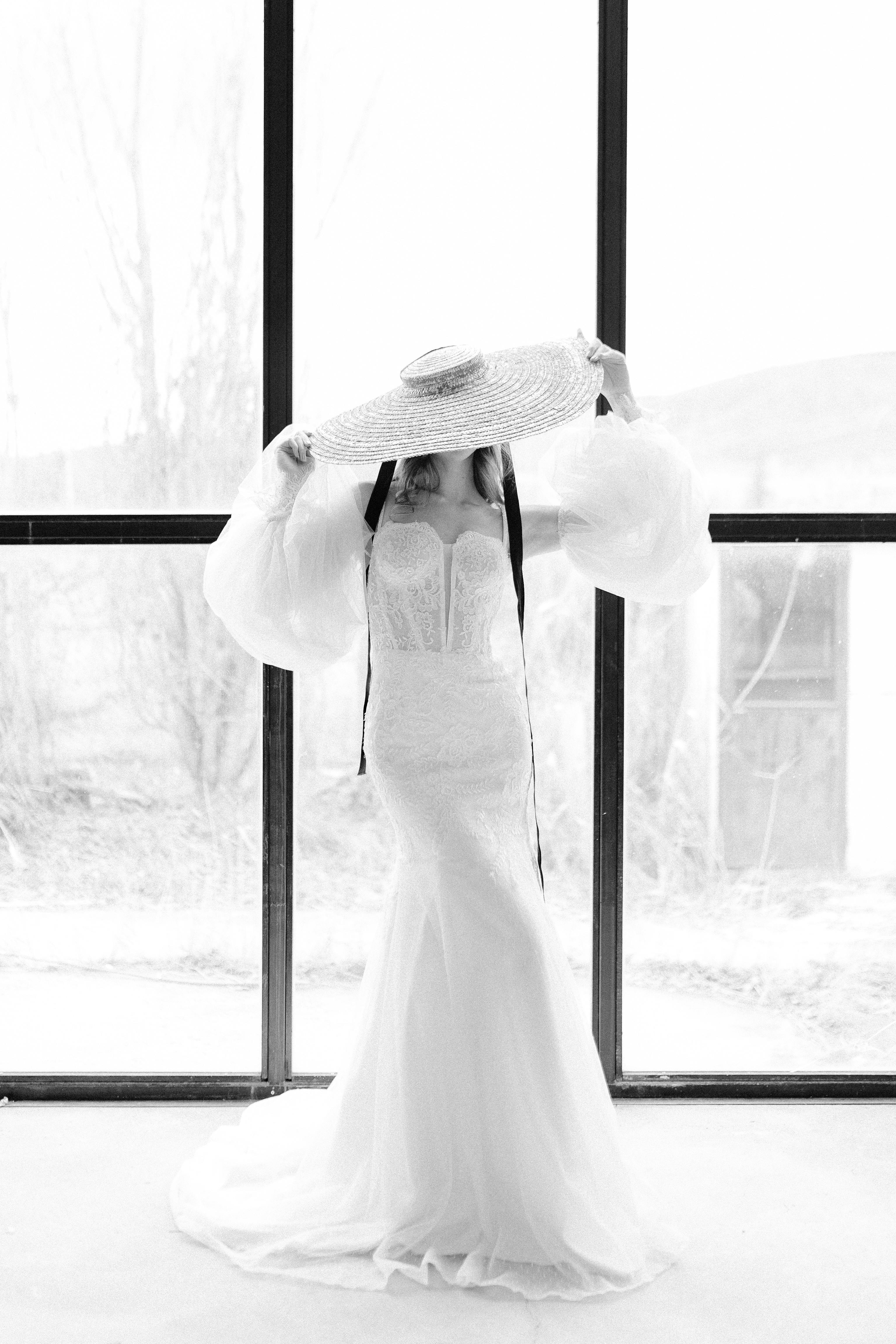 Chic black and white portrait of woman in elegant dress and wide-brimmed hat by the window.