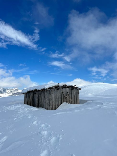 A wooden cabin set in a snowy landscape with blue skies and frosty mountains in the background.