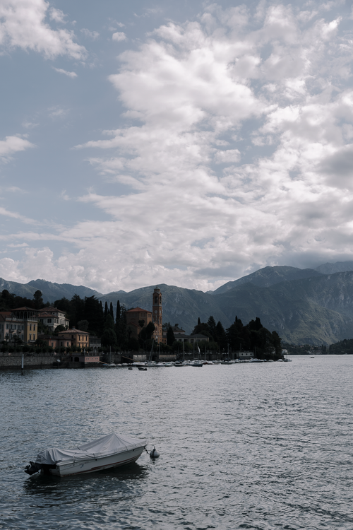 Free Peaceful scene of Lake Como with a boat, mountains, and tower under a cloudy sky. Stock Photo