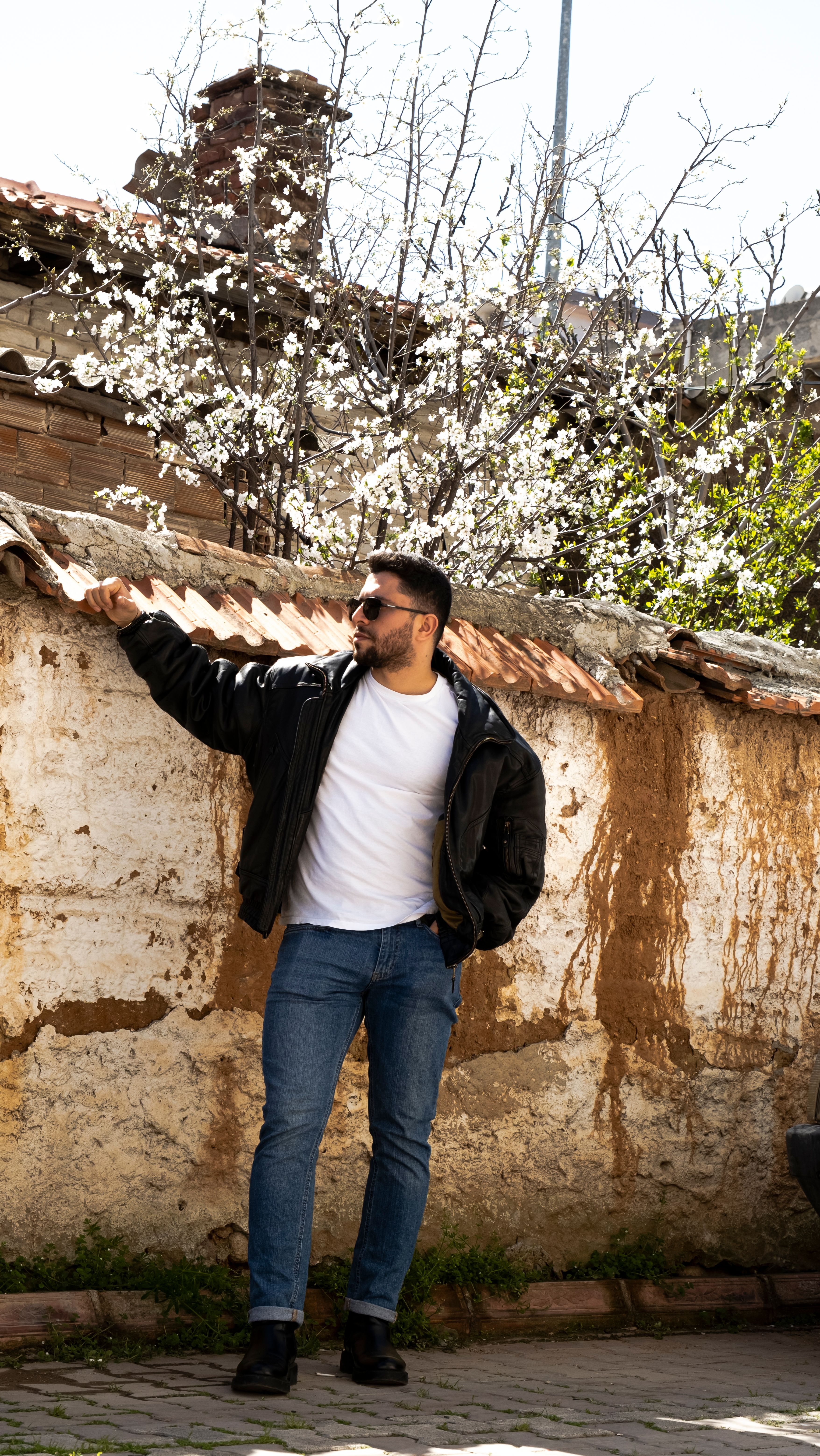 man in a trendy casual outfit standing next to a wall and a tree with white flowers